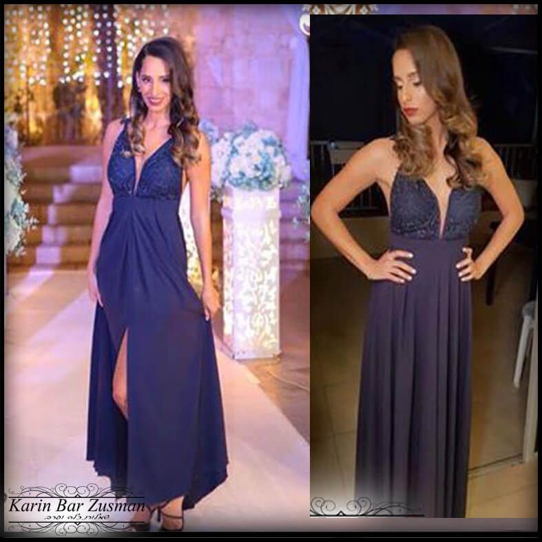 אישה בלבוש רשמי כחול כהה, עומדת בבית, מחייכת. שיער ארוך וגלי, מחוך תחרה, שס גבוה, סביבה מוארת היטב.