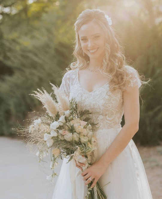 כלה בשמלת תחרה לבנה מחייכת, אוחזת זר. אור שמש רך, סביבה חיצונית.