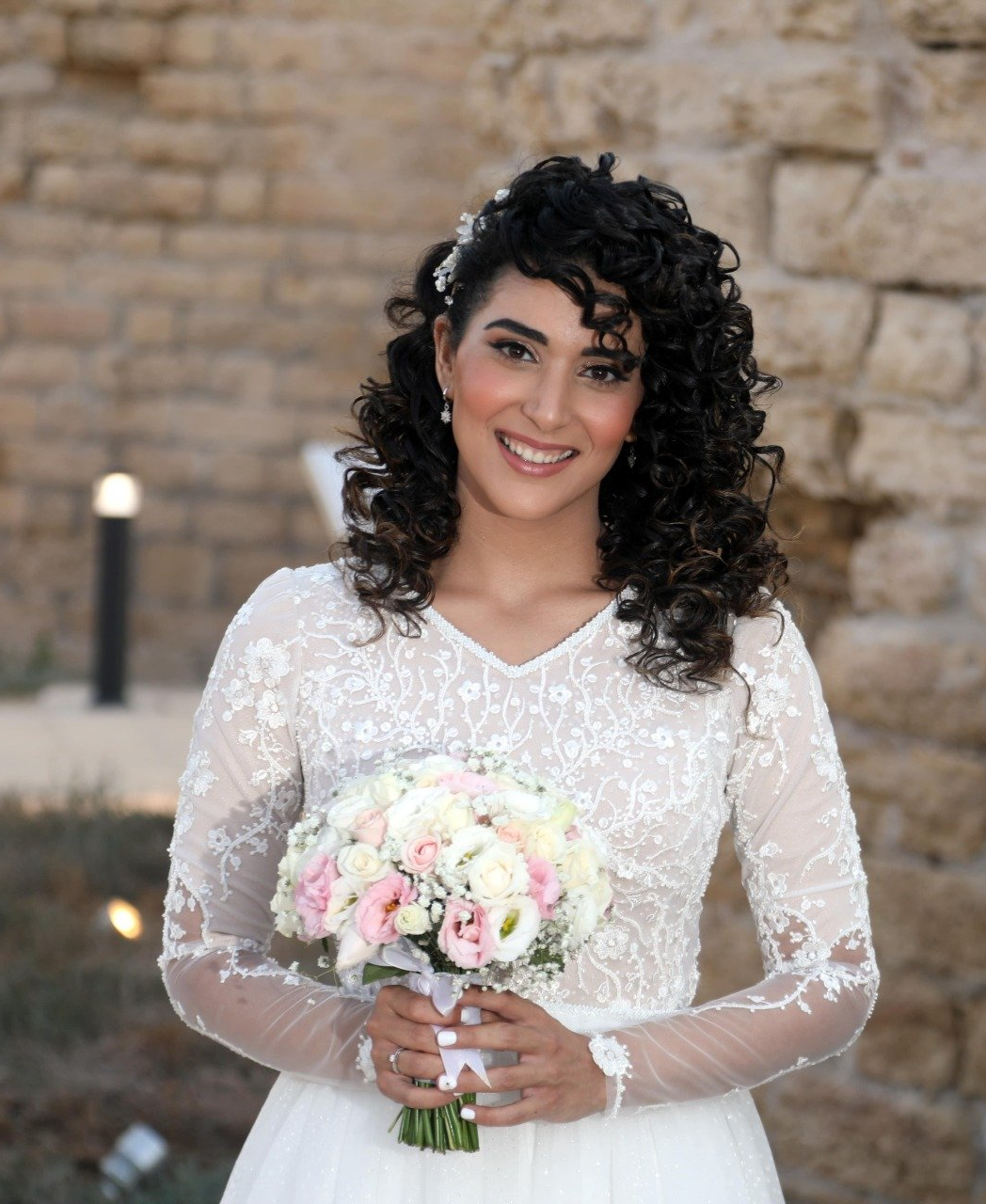 כלה בשמלת תחרה, אוחזת זר פרחים, מחייכת. שיער כהה מתולתל, רקע קיר אבן.