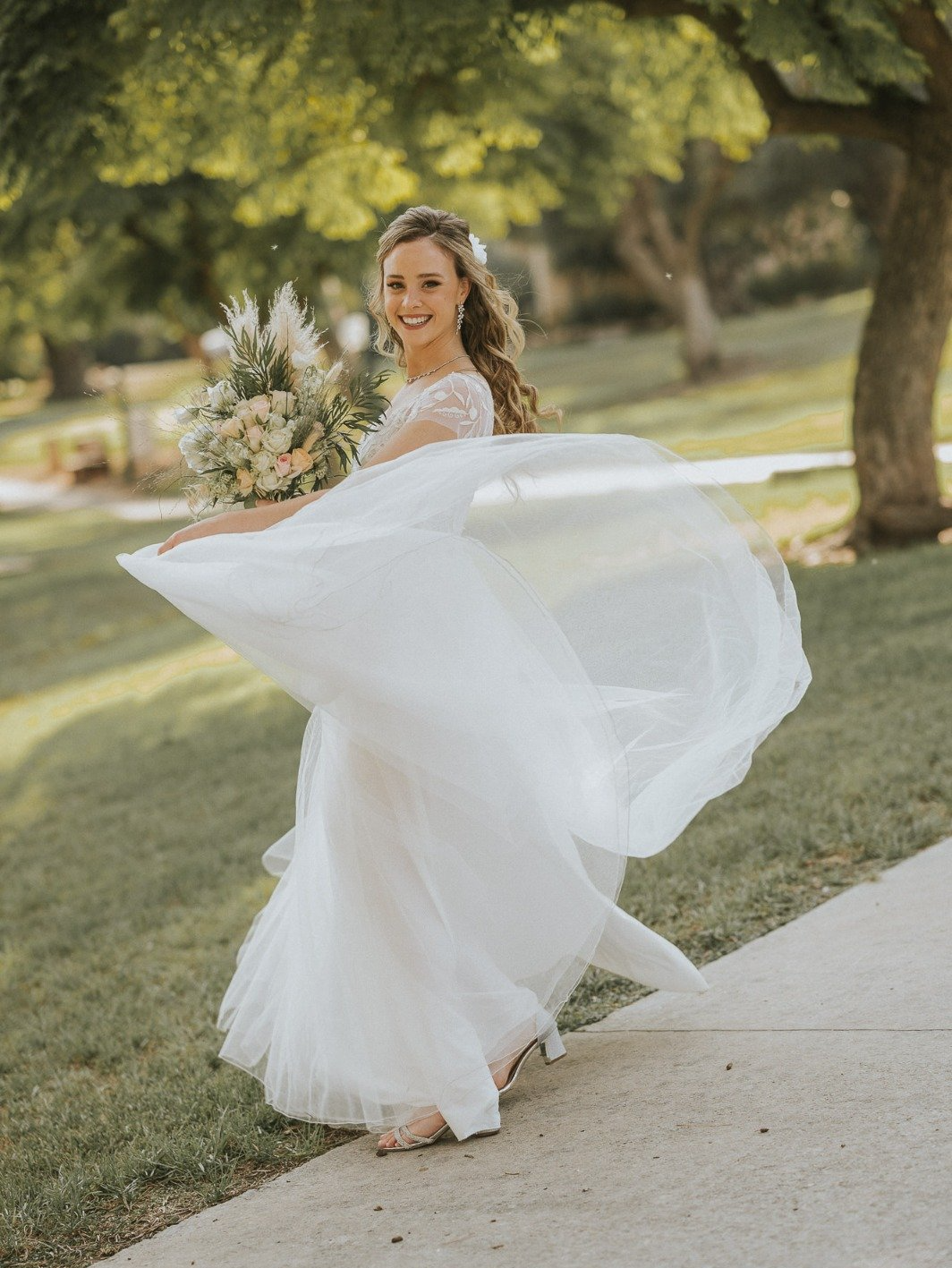 כלה בשמלה לבנה מסתובבת, מחייכת, אוחזת בפרחים על שביל. עצים ירוקים ודשא ברקע.