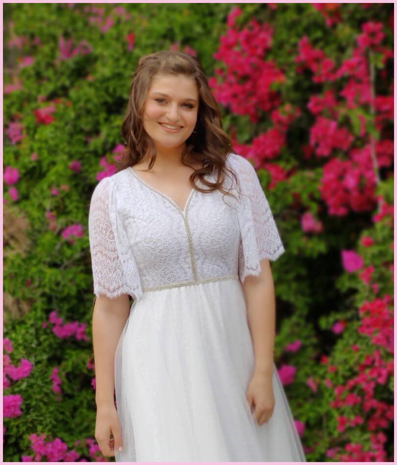 אישה בשמלה לבנה מחייכת, מצטלמת מול פרחים ורודים.