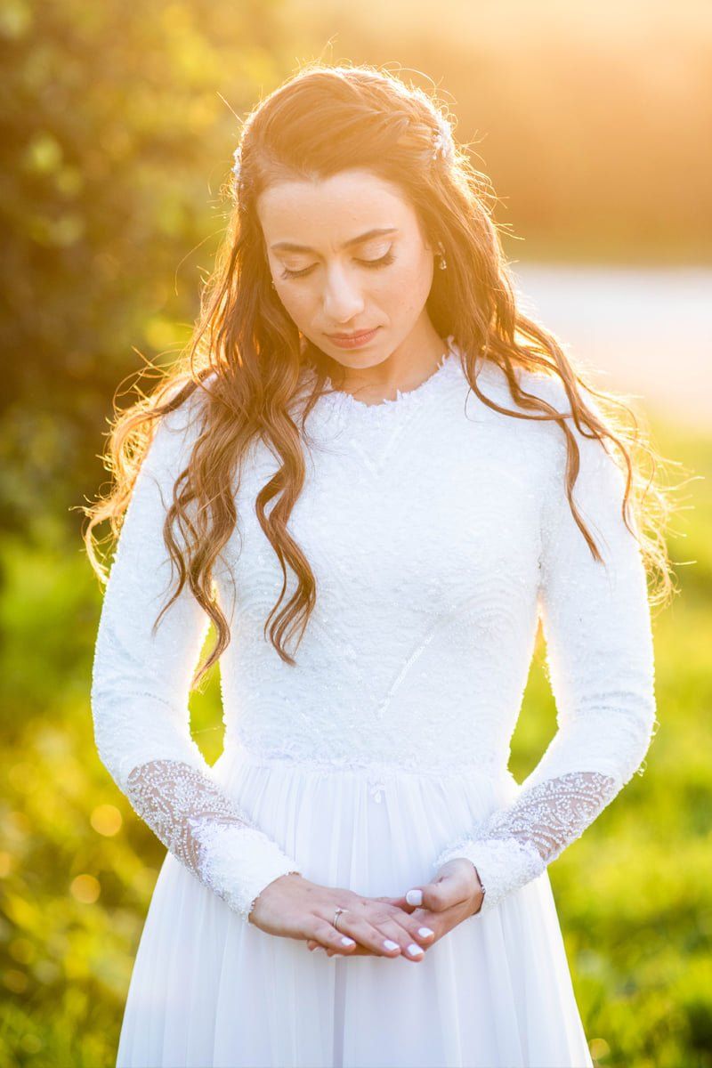 אישה בשמלה לבנה עם שיער ארוך, ידיים שלובות, בחוץ באור שמש זהוב.