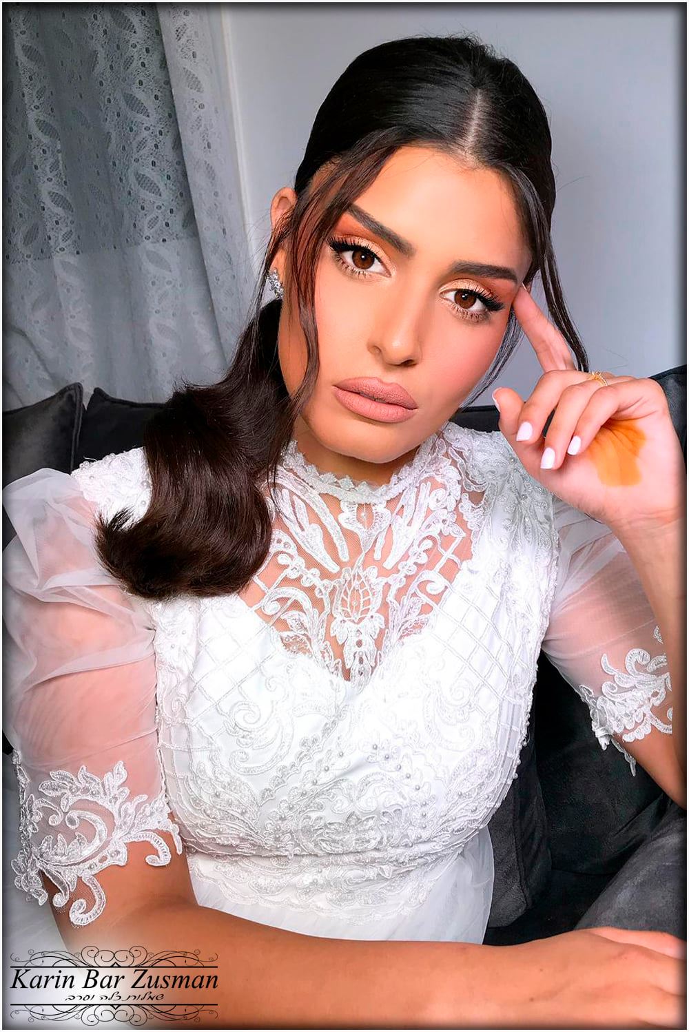 אישה בשמלת תחרה לבנה עם תספורת, מניחה את סנטרה ומביטה במצלמה במבט מעמיק.