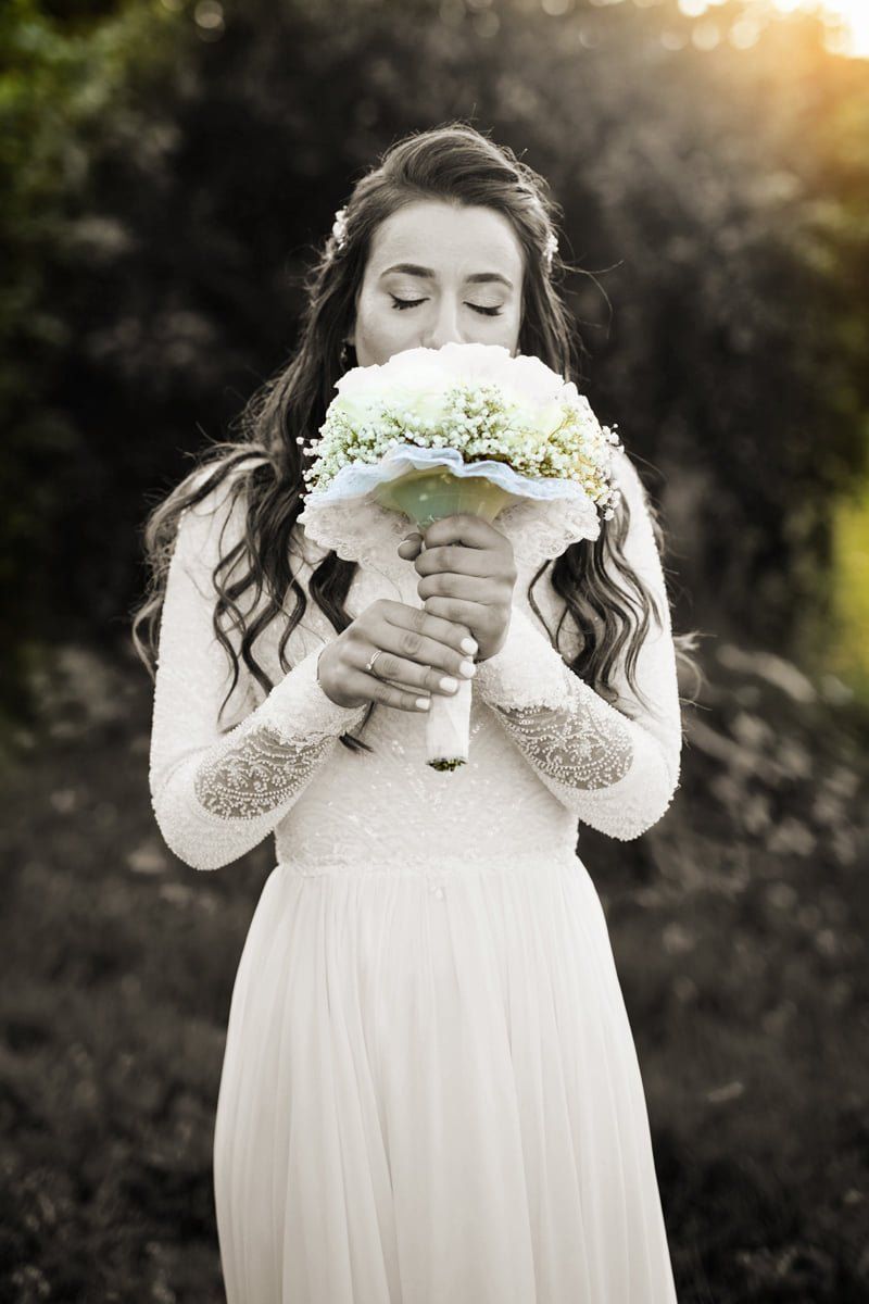 כלה אוחזת זר, מריחה פרחים, עיניים עצומות, לובשת שמלת תחרה ארוכת שרוולים בחוץ.