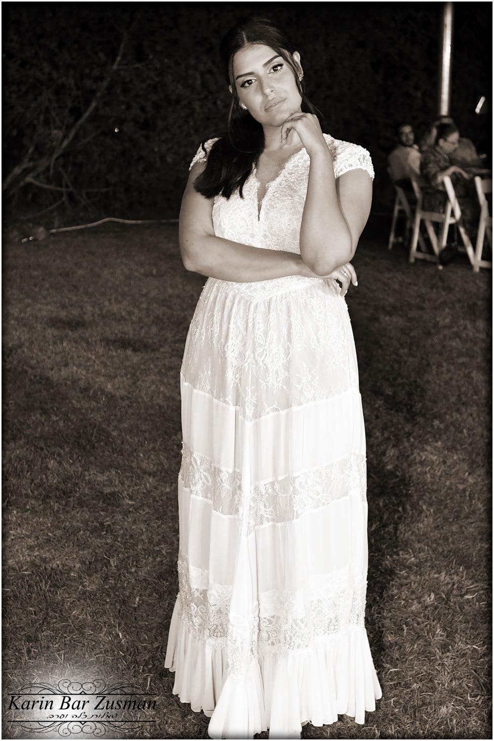 אישה בשמלת תחרה לבנה מצטלמת בחוץ, יד על הסנטר. שקיעת לילה, גוון ספיה.