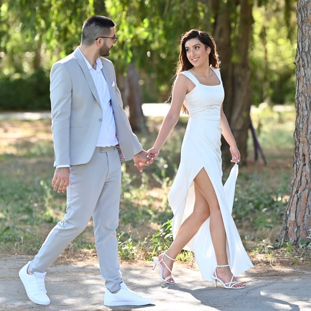 זוג הולך יד ביד בחוץ; גבר בחליפה, אישה בשמלה לבנה עם שס גבוה.