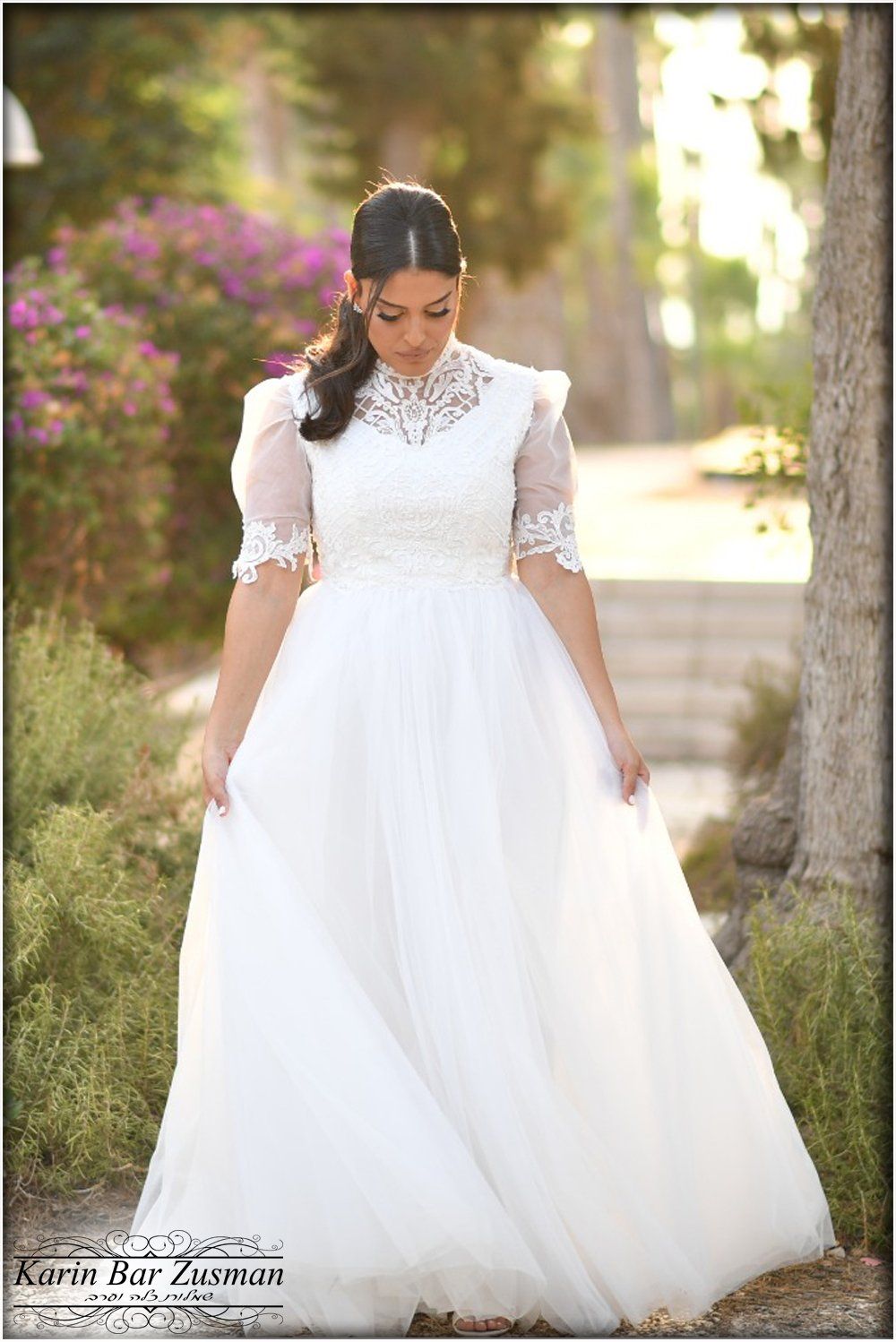 אישה בשמלת כלה לבנה, עומדת בחוץ. עלווה ירוקה שופעת ופרחים סגולים ברקע.