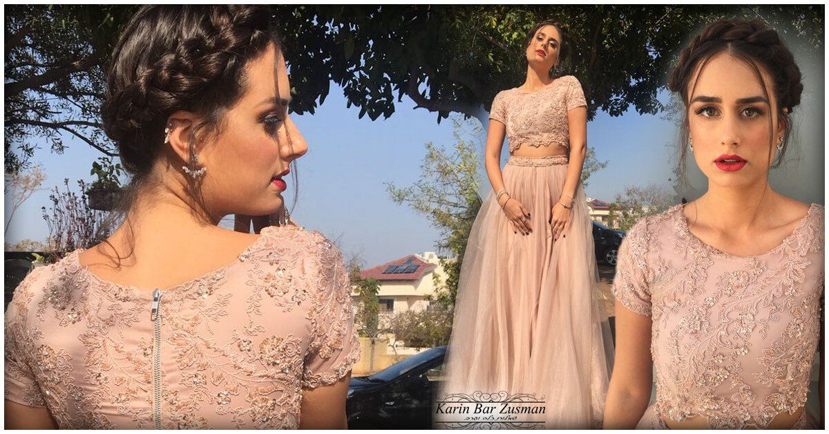 אישה בשמלה ורודה רקומה עם תסרוקת קלועה. מצטלמת בחוץ ליד עצים ובניין.