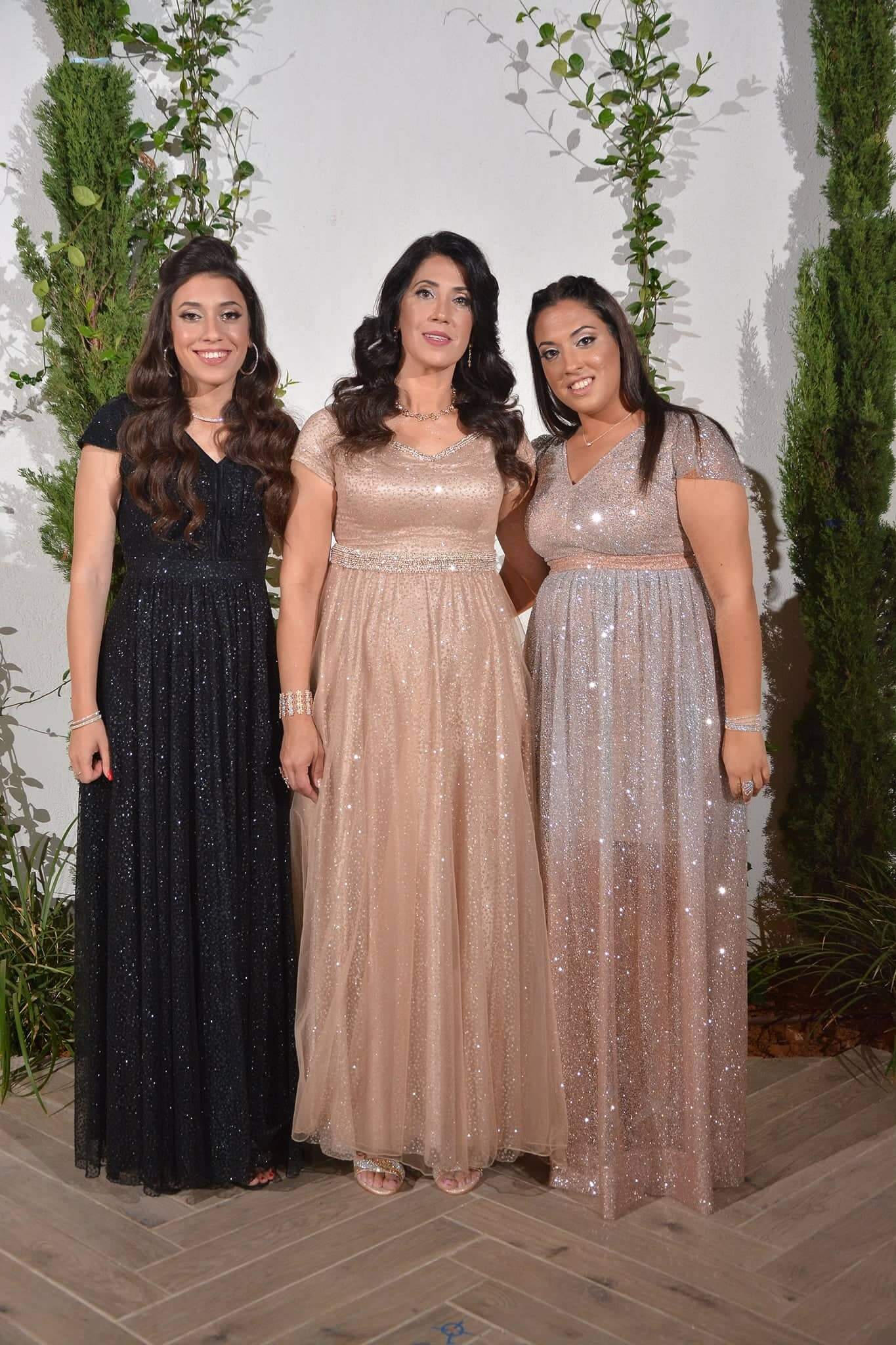 שלוש נשים בשמלות רשמיות מנצנצות, מחייכות, מצטלמות יחד, עומדות על רקע ירק.