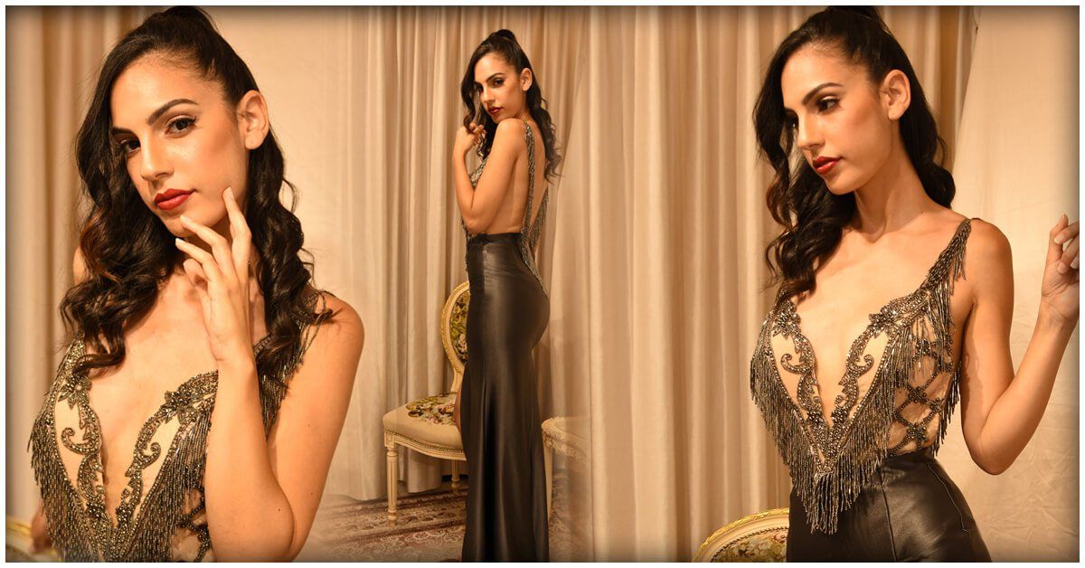 אישה בשמלה שחורה מעוטרת, מצטלמת בחדר עם וילונות בגוונים ניטרליים.