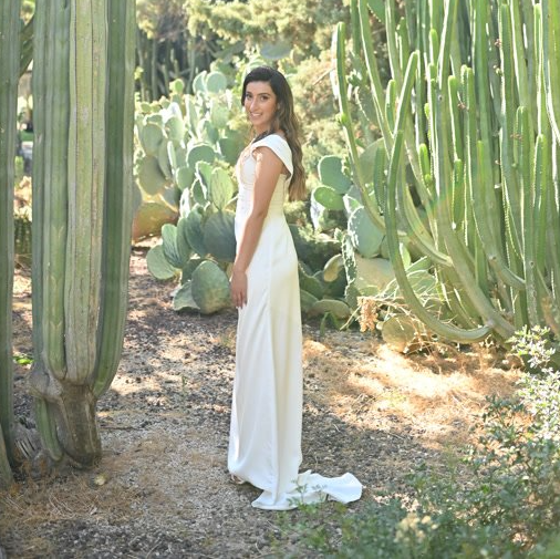 אישה בשמלה לבנה מצטלמת בגינה עם קקטוסים, מסתכלת מעבר לכתפה.