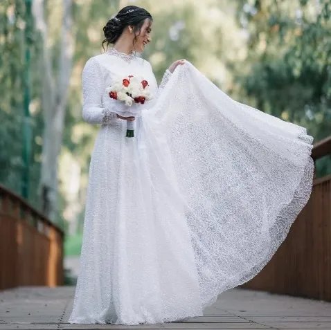 אישה בשמלת כלה לבנה אוחזת בפרחים על גשר בחוץ.