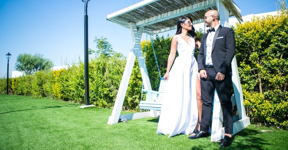 חתן וכלה בתנוחת גינה ליד נדנדה לבנה. הכלה לובשת שמלה לבנה, החתן בחליפה שחורה, שניהם מחייכים.