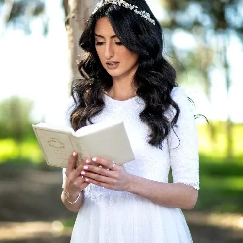 אישה בשמלה לבנה וכיסוי ראש, קוראת ספר בחוץ.