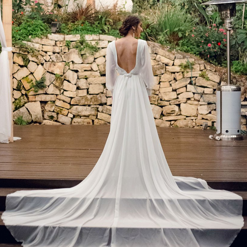 כלה בשמלת כלה לבנה עם שובל ארוך, עומדת על במה מעץ על רקע קיר אבן.