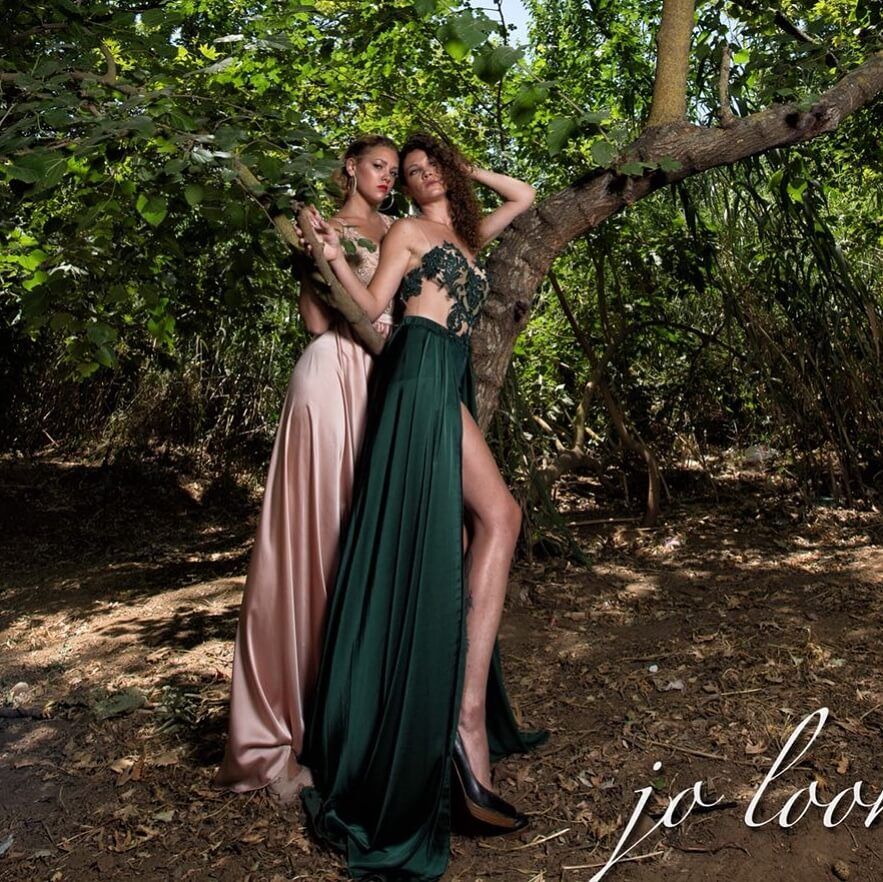 שתי נשים בשמלות אלגנטיות מצטלמות ליד עץ באזור מיוער. אחת לובשת ורוד, השנייה ירוק.