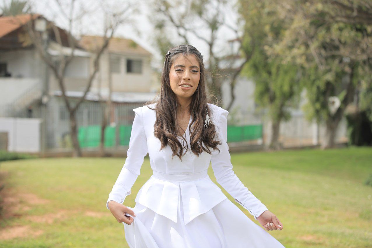 אישה בשמלה לבנה בחוץ, מחייכת, ידיים על חצאית, מדשאה ירוקה, רקע מטושטש.