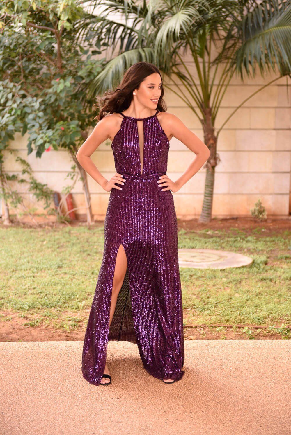 אישה בשמלה סגולה נוצצת עם שס, ידיים על הירכיים, בחוץ.