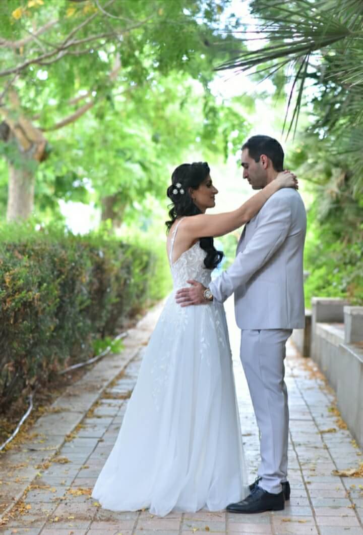 חתן וכלה מתחבקים בחוץ על שביל אבן, מוקפים בירק. היא לובשת שמלה לבנה, הוא לובש חליפה בהירה.