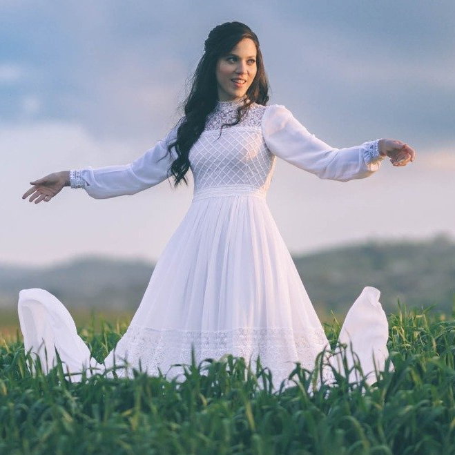 אישה בשמלה לבנה, ידיים מושטות קדימה, עומדת בשדה ירוק בשקיעה.