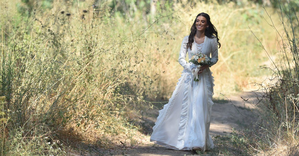 אישה בשמלת כלה לבנה מחייכת, הולכת בשביל עפר דרך דשא צהוב גבוה.