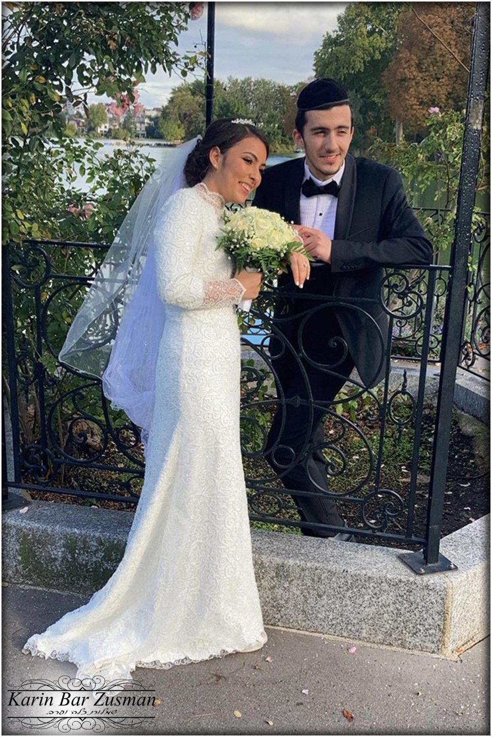 חתן וכלה מצטלמים בחוץ ביום חתונתם. כלה בשמלה לבנה, אוחזת פרחים, מחייכת. חתן בטוקסידו שחור.