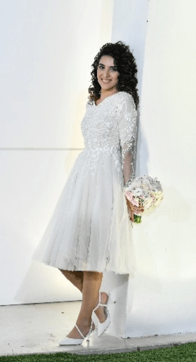 אישה בשמלת כלה לבנה באורך הברך, אוחזת זר פרחים, נשענת על קיר לבן, מחייכת.