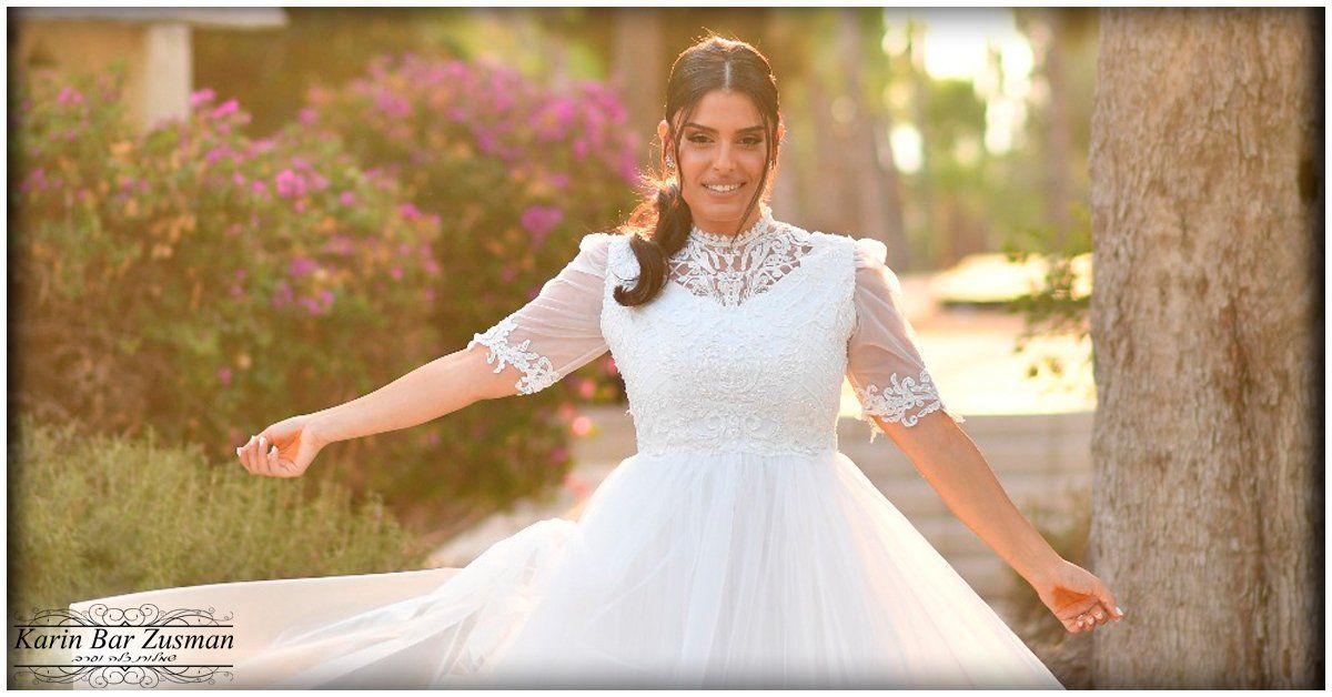 אישה בשמלת כלה לבנה, ידיים מושטות, מחייכת בחוץ ליד שיחים.