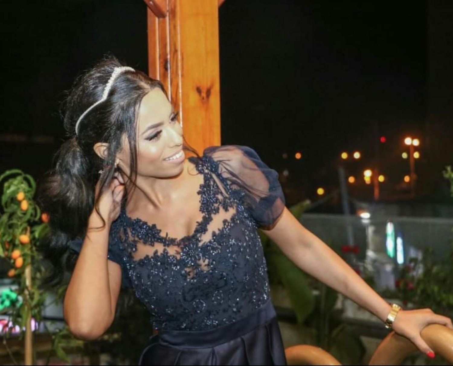 אישה בשמלה כחולה כהה, מחייכת עם שיער כהה אסוף בקוקו, נשענת ליד מבנה עץ בלילה.