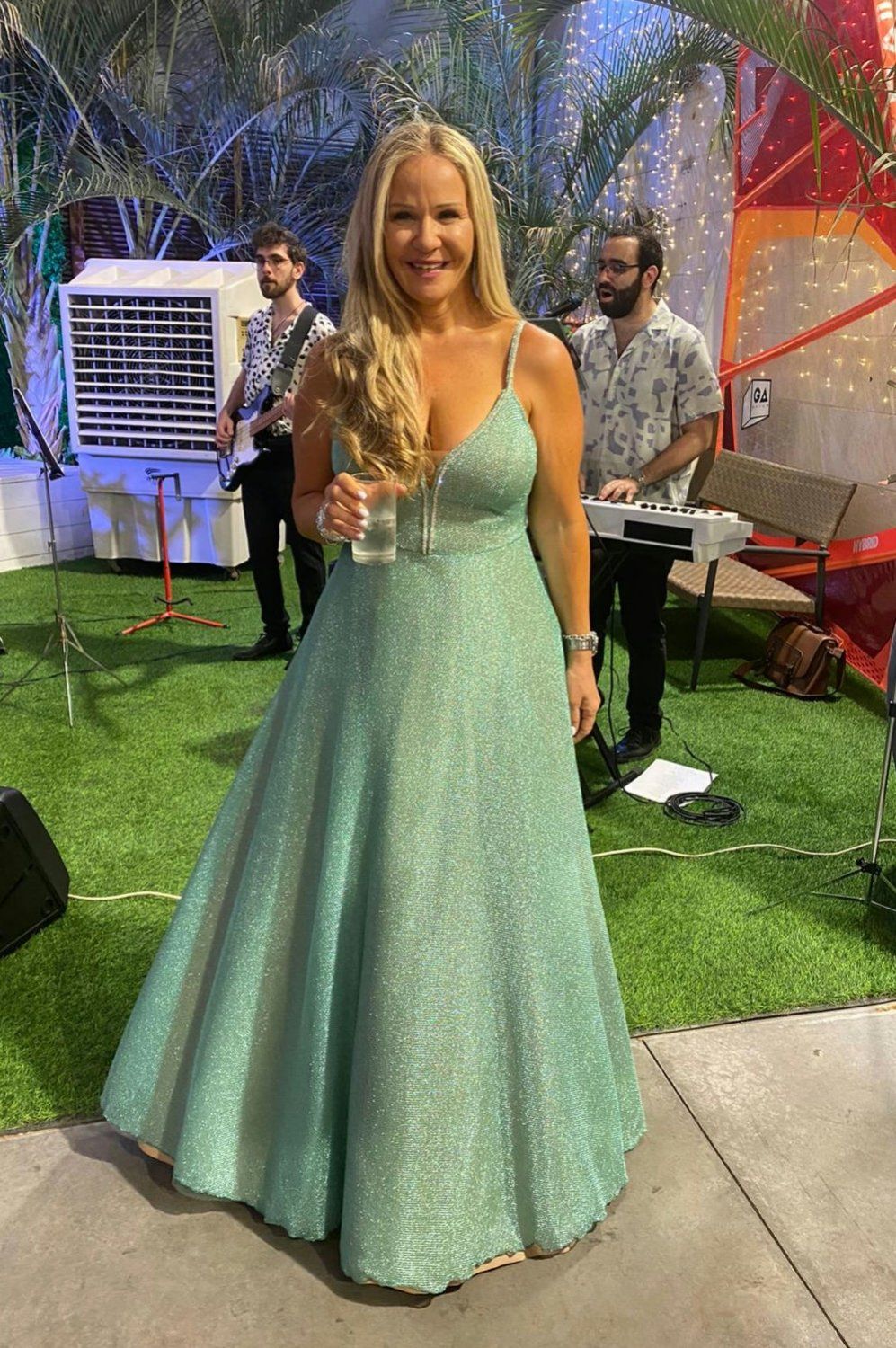 אישה בשמלה בצבע טורקיז נוצץ אוחזת כוס, מחייכת במסיבה עם להקה ברקע.