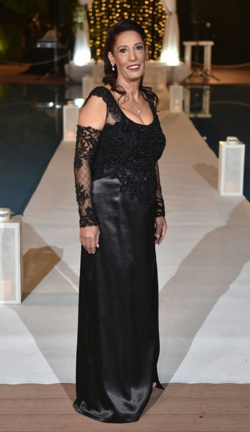 אישה בשמלת ערב שחורה עומדת על שביל, מחייכת. תפאורה חיצונית עם שרשרת אורות ומים.