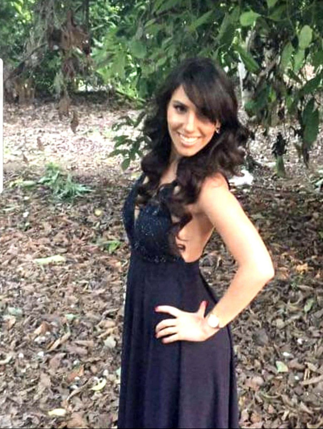 אישה בשמלה כחולה כחולה מצטלמת בחוץ עם חיוך, ידיים על הירכיים, מול עצים עלים.