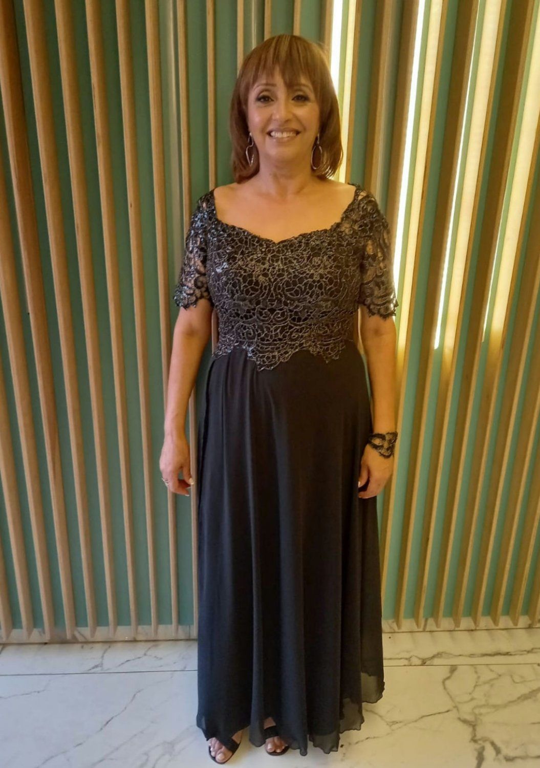 אישה עם שיער חום קצר, לובשת שמלה שחורה רשמית, מחייכת, מול קיר מחוספס.