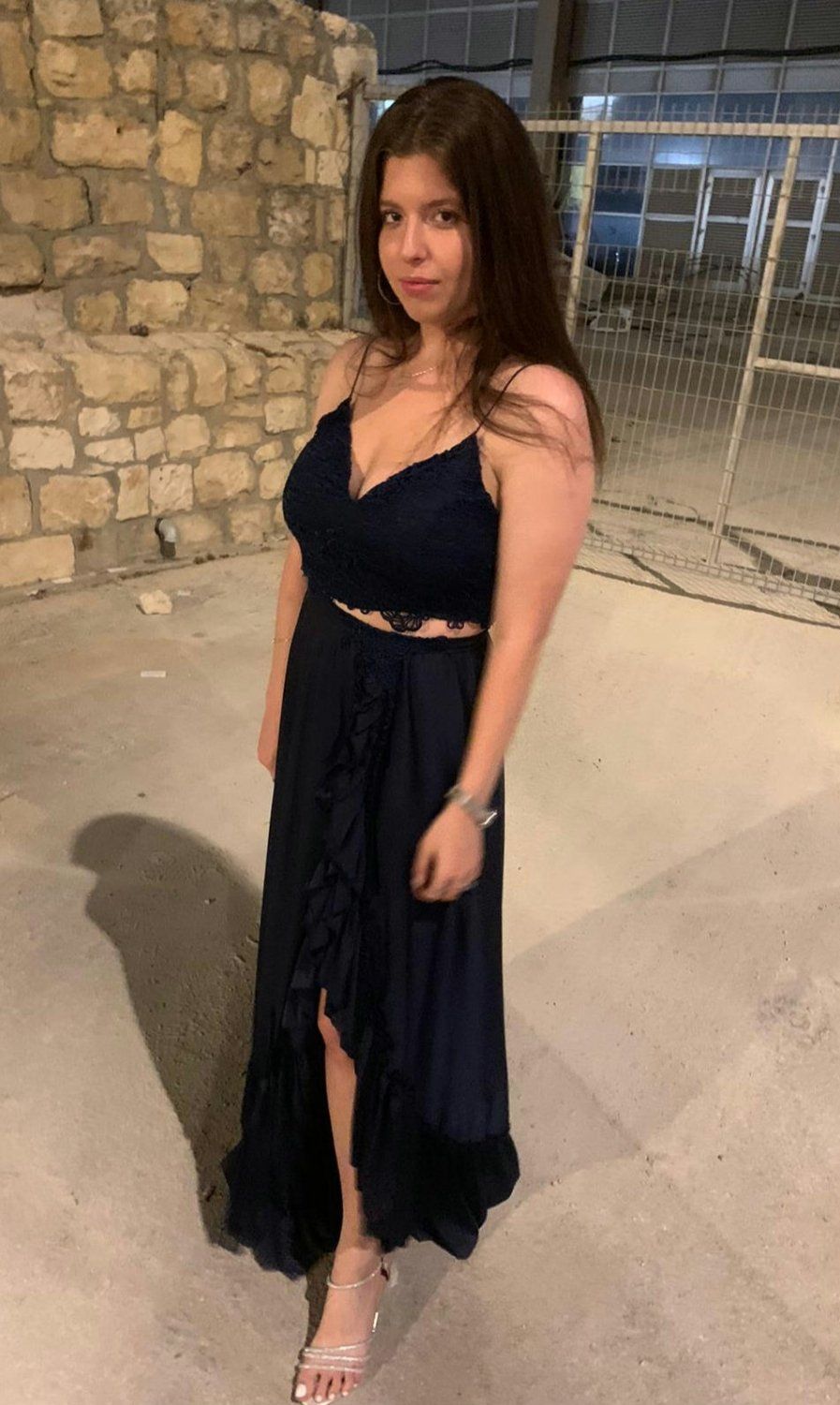 אישה בשמלה כחולה כהה עם שס גבוה, עומדת בחוץ ליד קיר אבן.