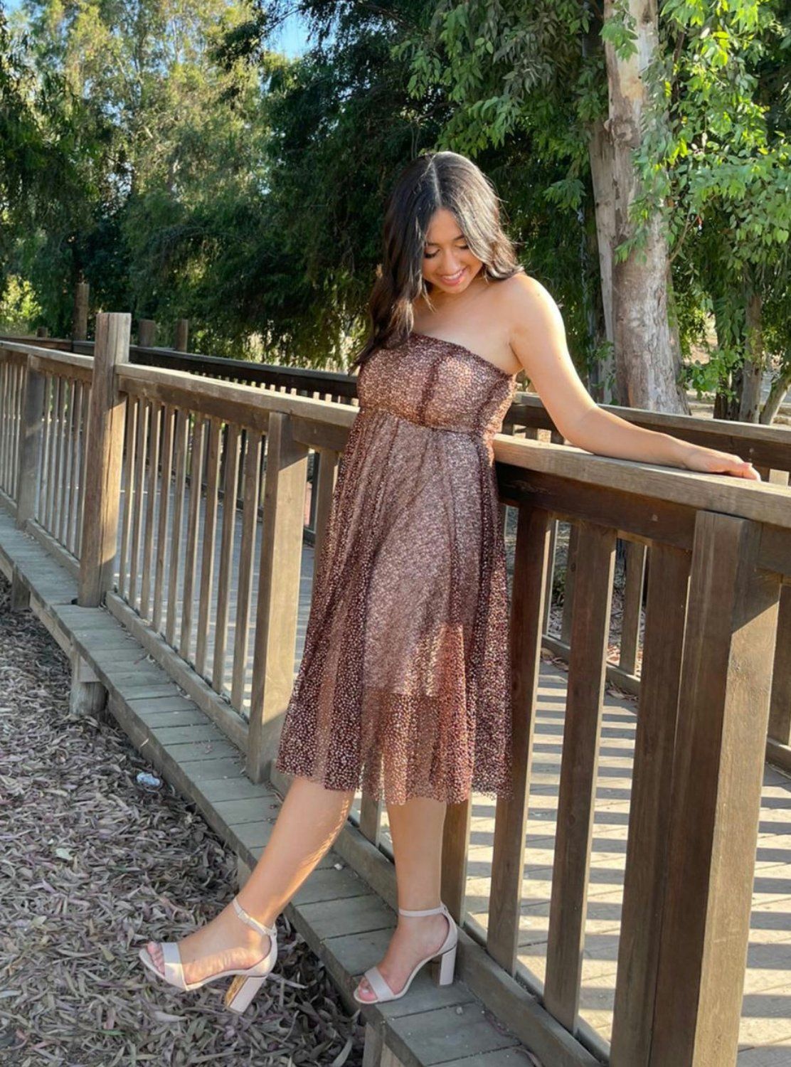 אישה בשמלה נוצצת, נשענת על גשר עץ בפארק חיצוני, מחייכת.