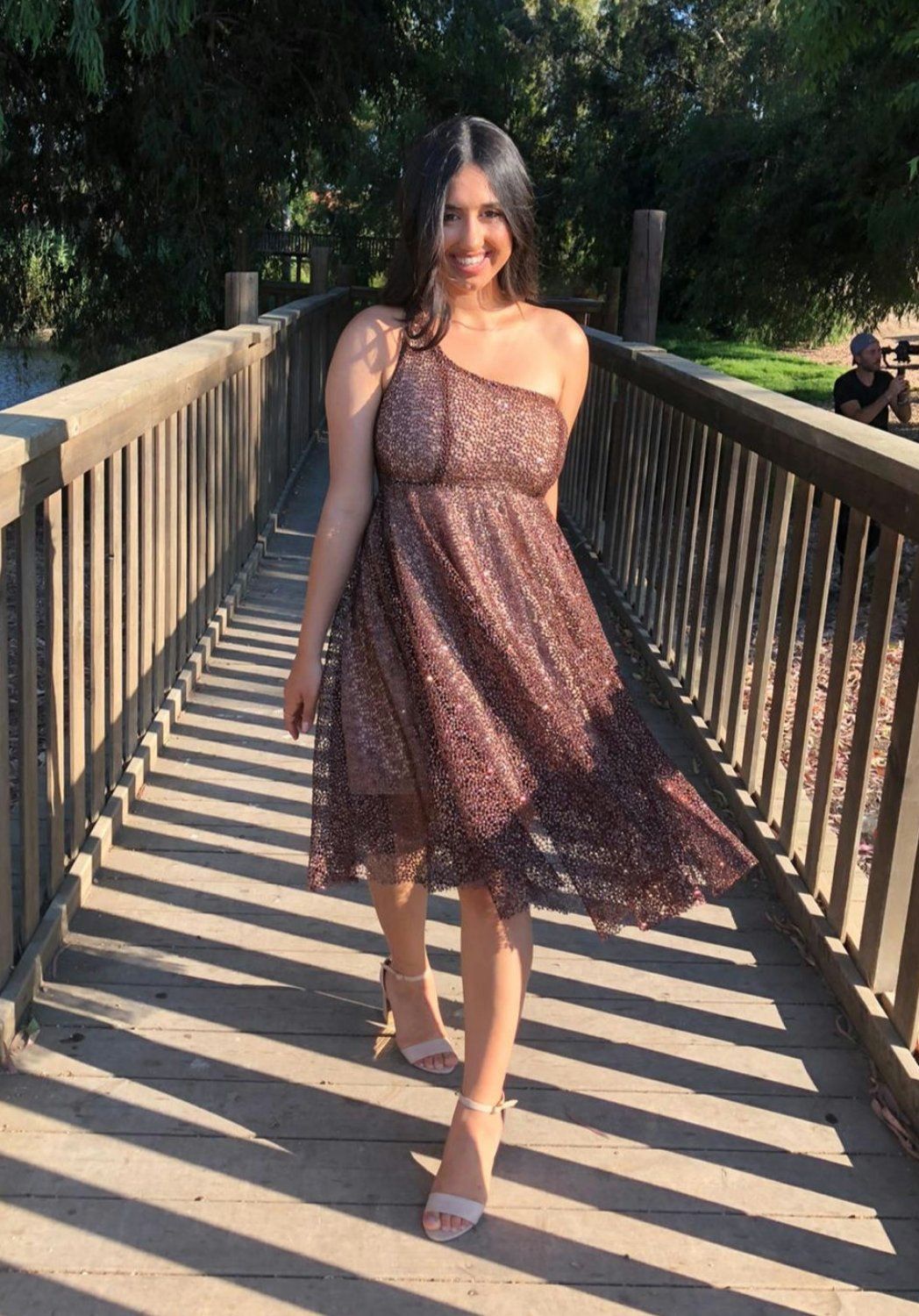 אישה בשמלה חומה עם כתף אחת הולכת לעבר הצופה על גשר עץ, מחייכת, בחוץ.