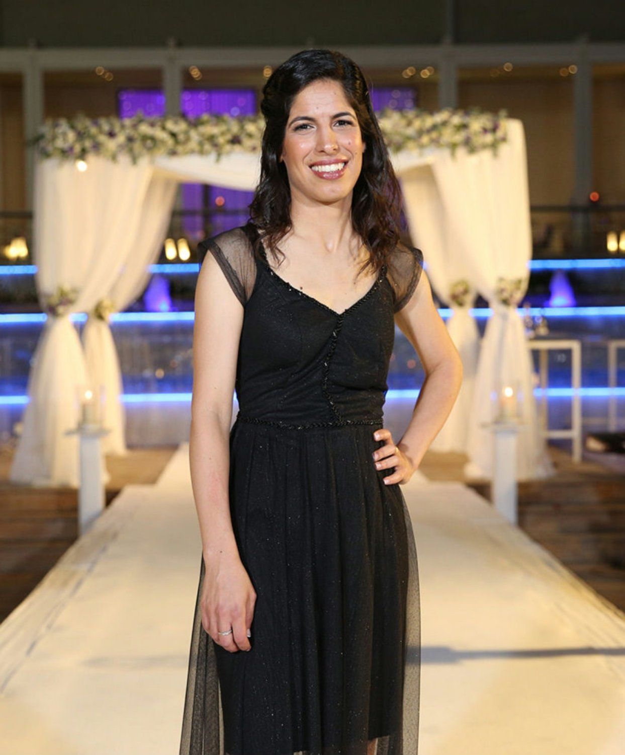 אישה בשמלה שחורה מחייכת, מצטלמת מול רקע טקס חתונה עם קישוטים לבנים וכחולים.