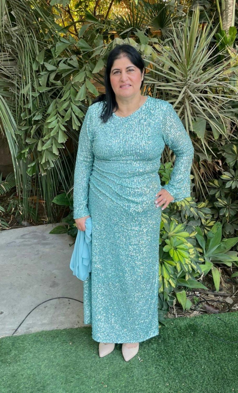 אישה בשמלה ארוכה, טורקיז, משובצת פאייטים מצטלמת בחוץ. היא אוחזת בארנק, מחייכת ונועלת נעלי עקב בצבע גוף.