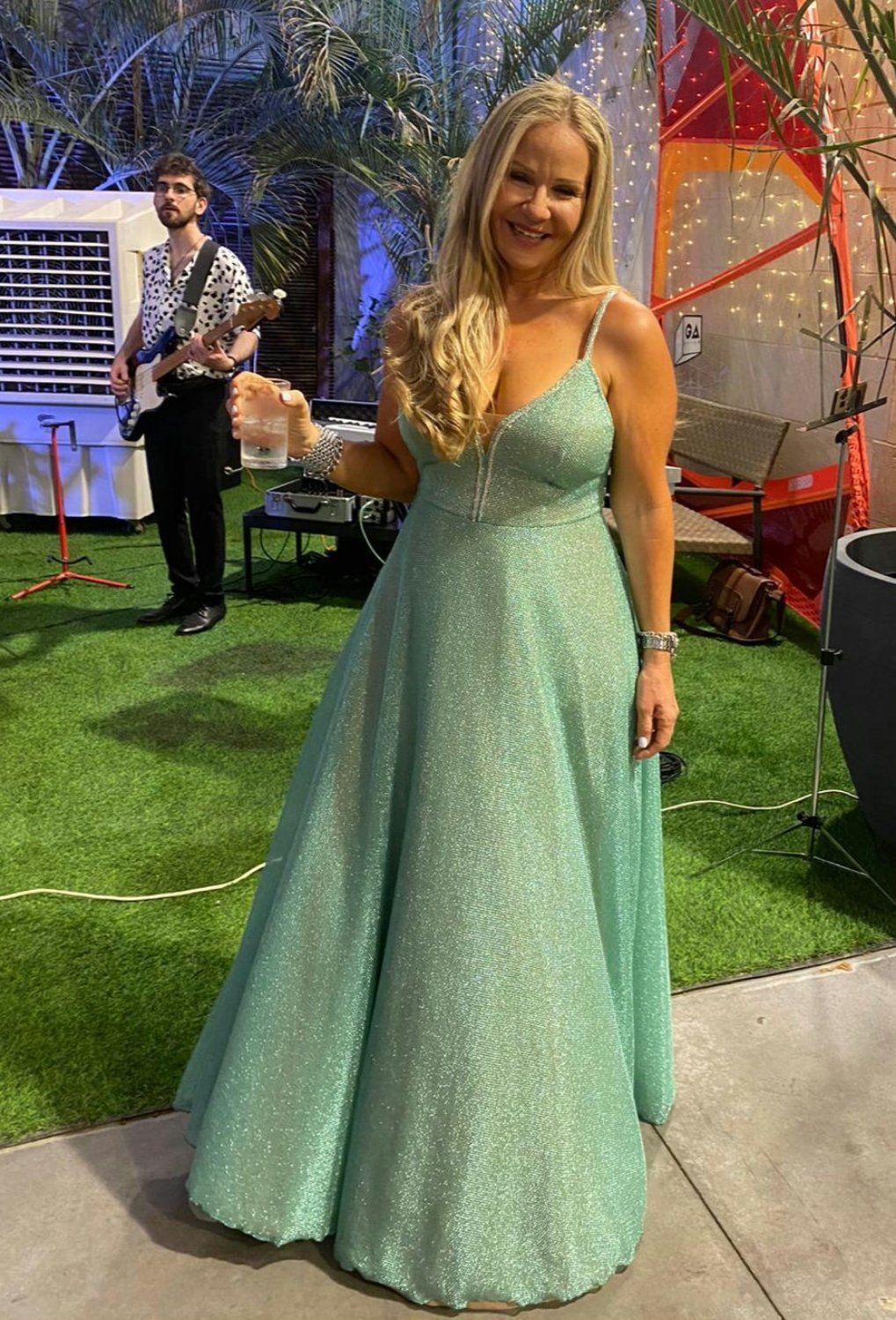 אישה בשמלה ירוקה נוצצת, מחייכת, באירוע חיצוני. חבר להקה ברקע.