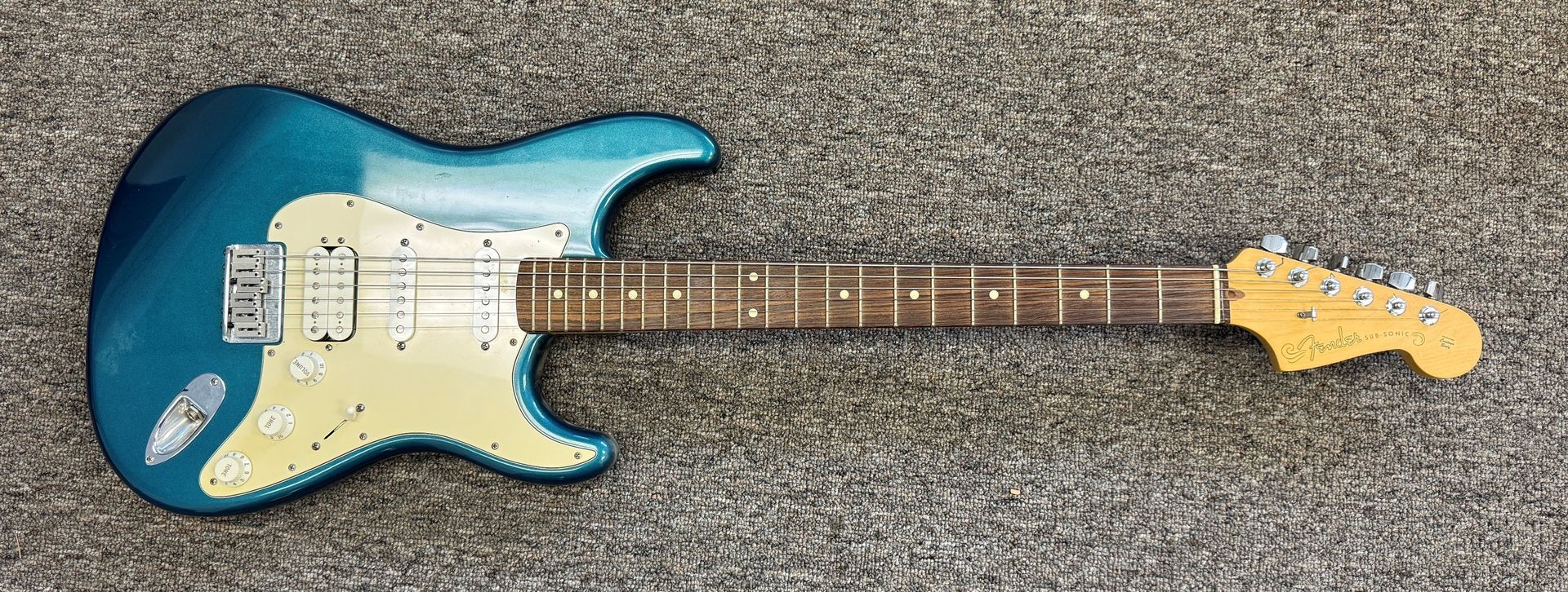 Blue electric guitar lying on a gray carpet, with a white pickguard and maple neck.