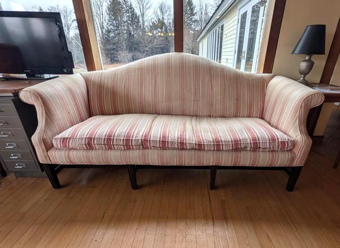 A camelback sofa upholstered in a light-colored, vertically striped fabric, centered in a room with hardwood floors.