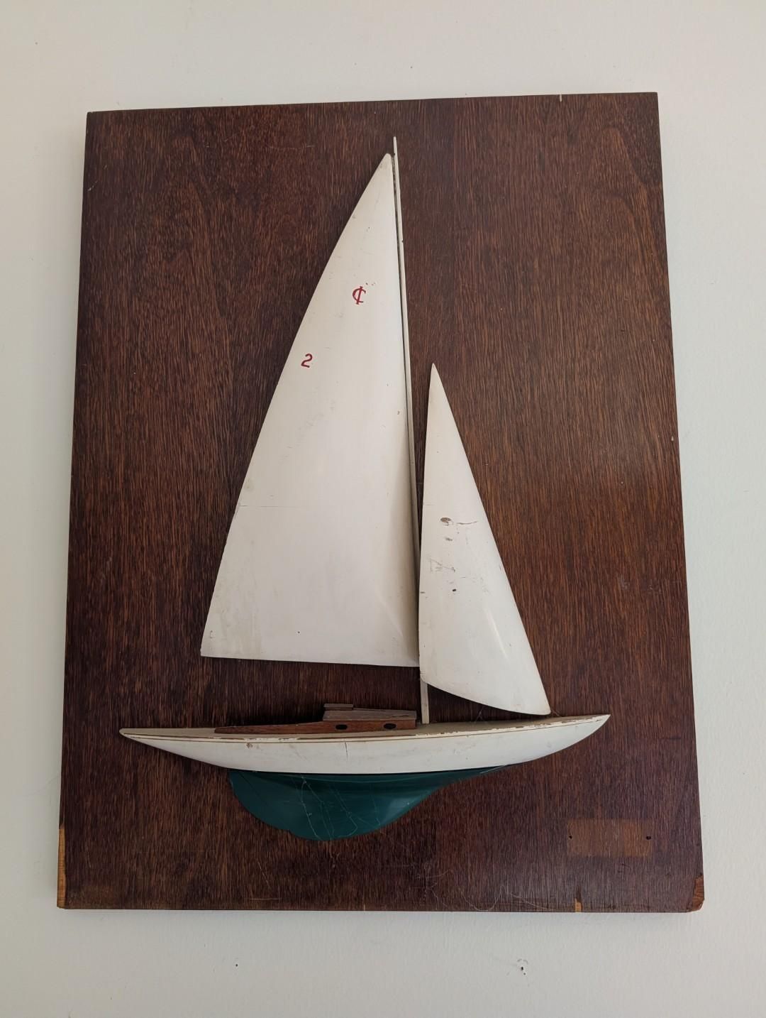 A 3D wooden wall art piece featuring a small white sailboat with two sails mounted on a dark brown wooden rectangular base.