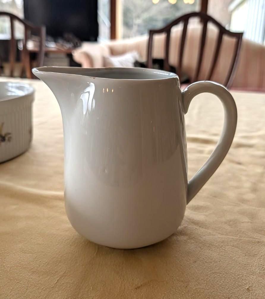 A plain white ceramic creamer pitcher sits on a light-colored tablecloth, with a partial view of a dining room behind.