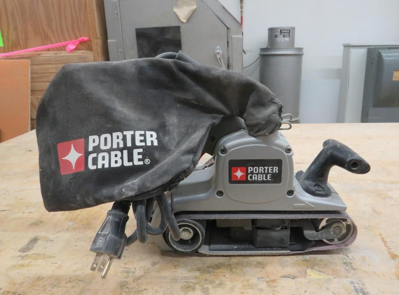 Gray Porter-Cable belt sander with dust bag and power cord on a wooden surface.