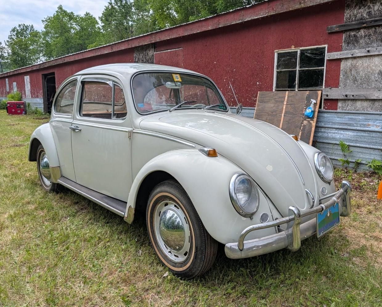 Vintage white Volkswagen Beetle parked beside a red barn on grass.