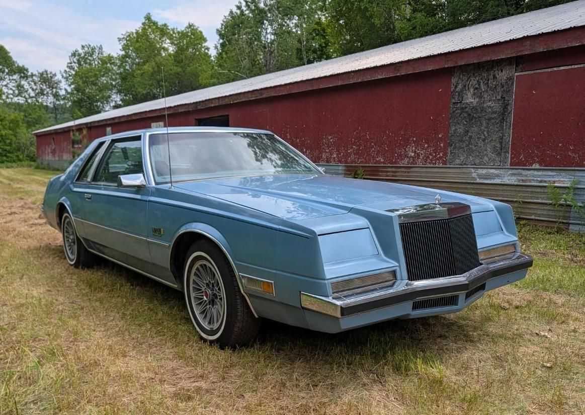 Blue vintage sedan parked on grass beside a red barn and trees