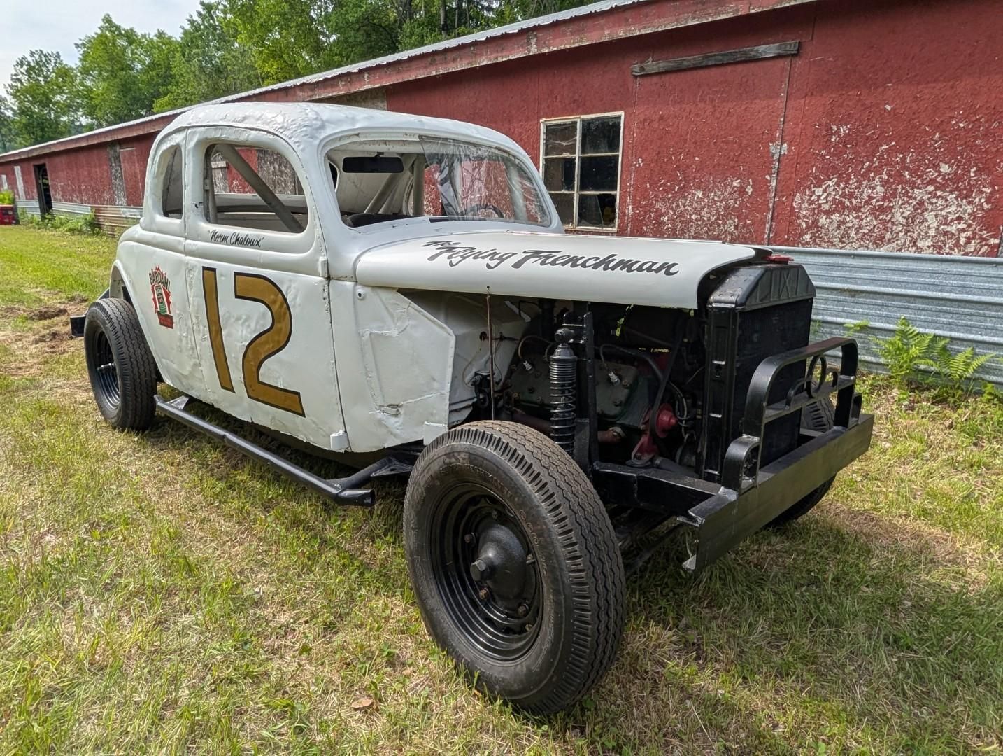 White vintage race car numbered 12 parked beside a red barn, front end exposed.
