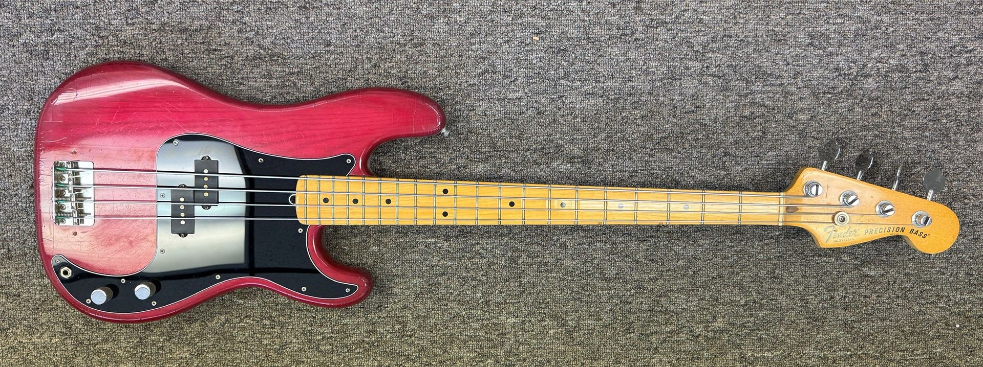 Red electric bass guitar with a black pickguard lying on a carpeted floor.