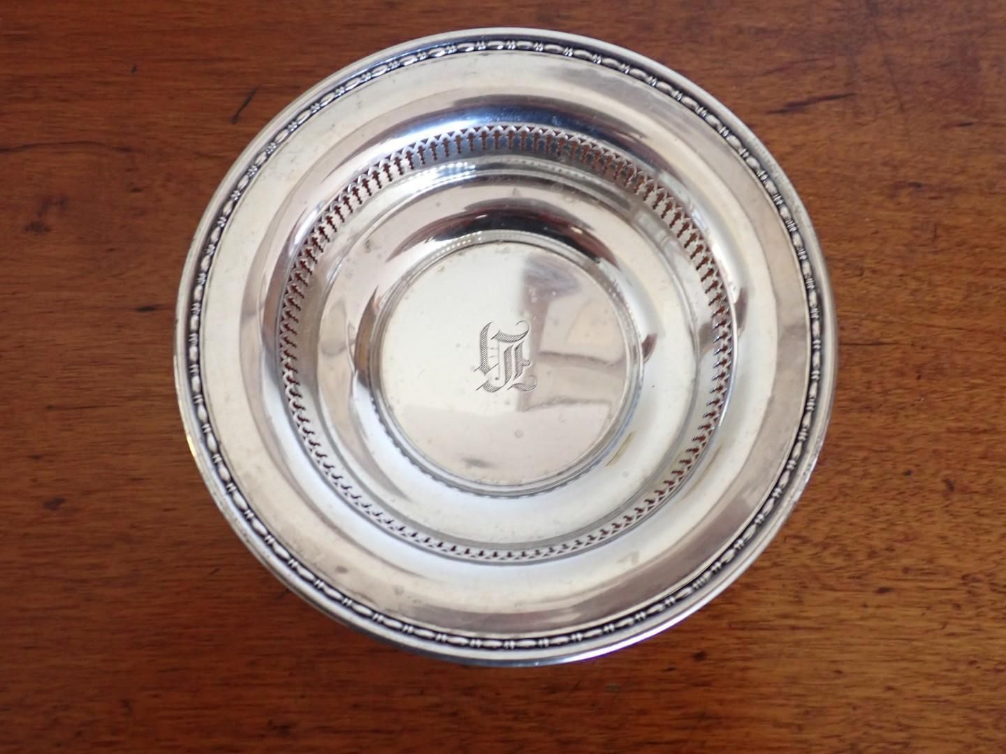 A silver-colored metal bowl with an embossed decorative rim and an engraved monogram in the center, resting on a wood table.