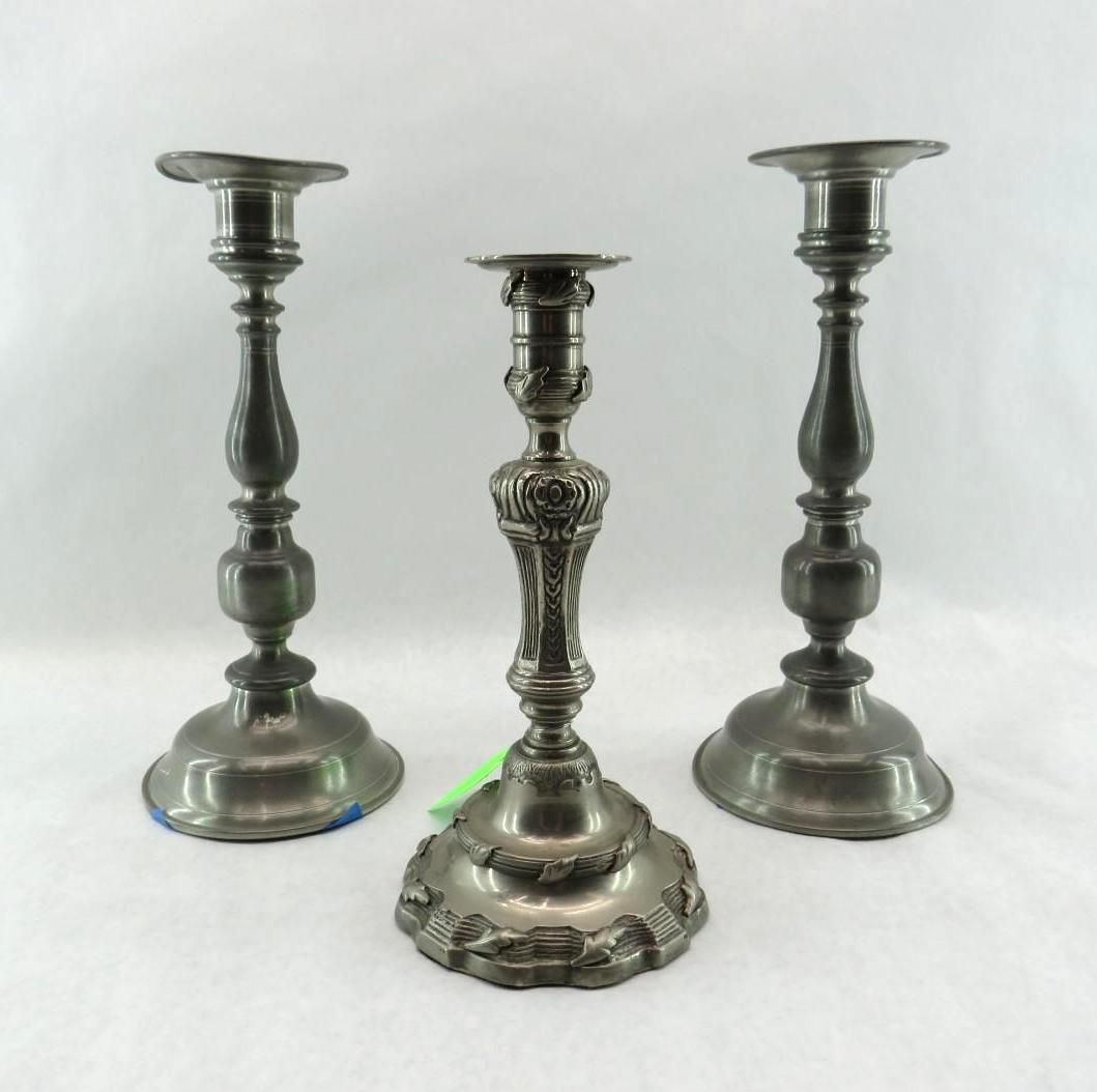 Three silver candlesticks on a white surface. One has ornate carvings.